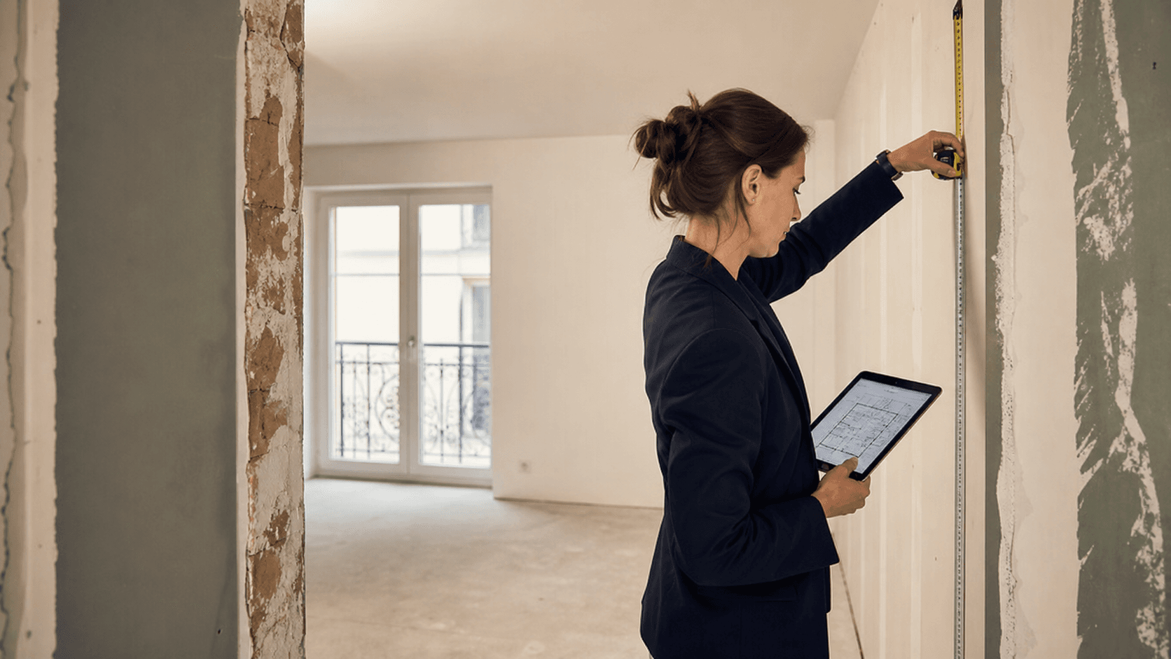Professionnelle mesurant une paroi sur un chantier de rénovation avec un plan en main